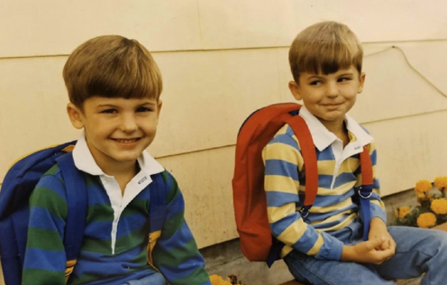 The Allred brothers as kids, sitting side by side on a porch in striped rugby shirts with backpacks.